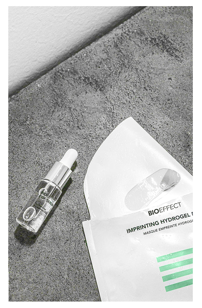 Black and white image of BIOEFFECT EGF Skincare Imprinting Hydrogel Face Mask coming out of the package and a bottle of EGF Serum laying on a concrete surface.